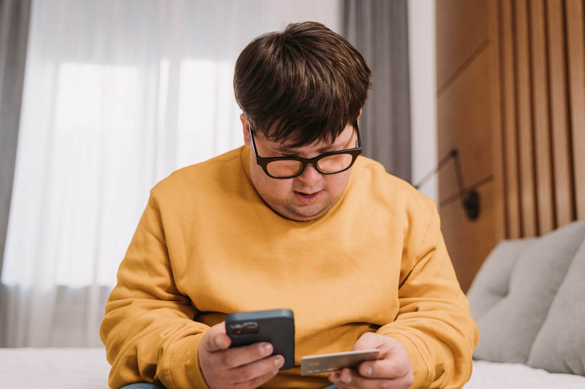 A person looks at a smartphone with a worried expression while holding a credit card, illustrating possible scam awareness.