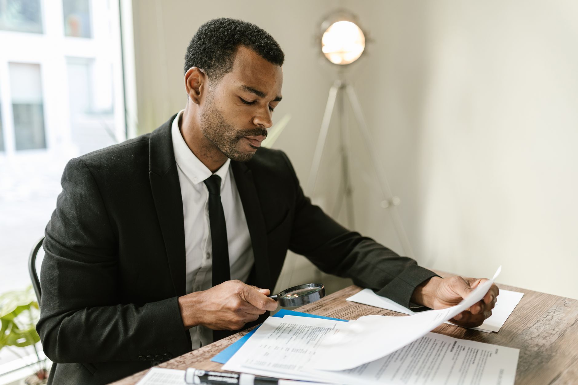 A person at a desk compares two paper documents with a magnifying glass while taking notes