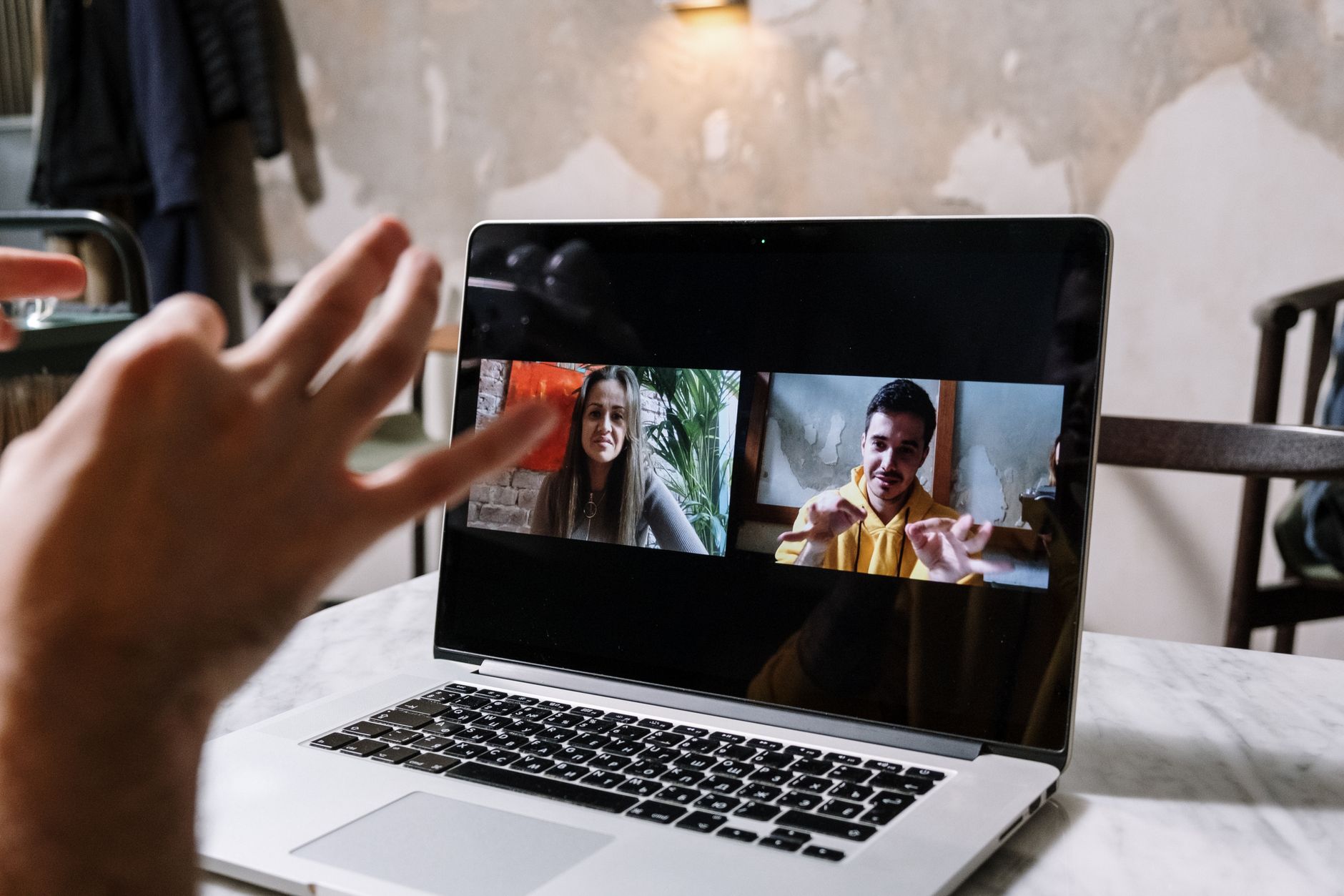 Two people on a laptop video call, one smiling and holding up three fingers to the camera.