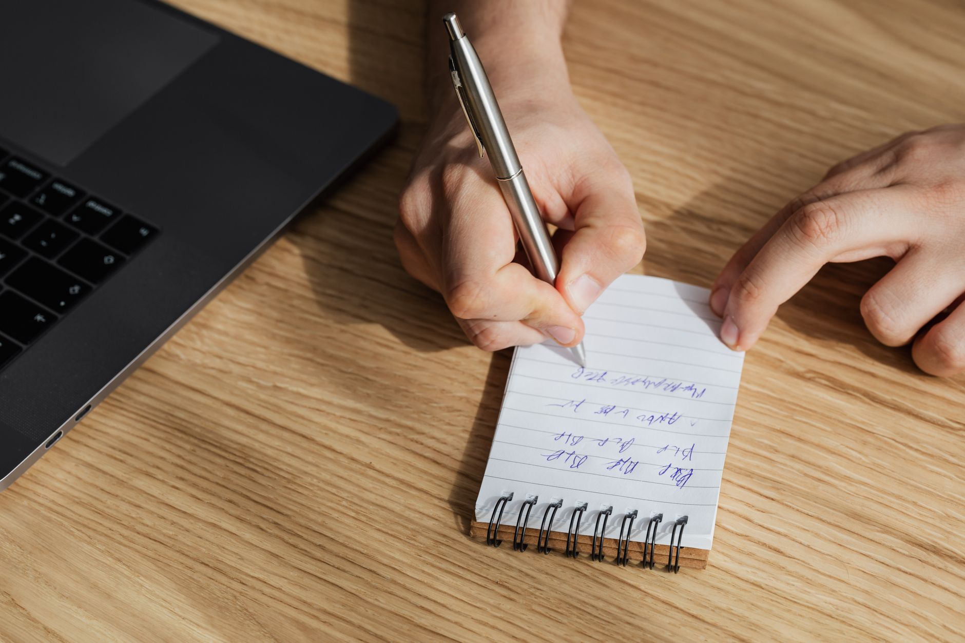 A person sitting at a kitchen table writing verification questions on a notepad next to a smartphone.
